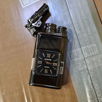 Briquet en métal avec horloge intégrée. Au-dessus du briquet, il y a un petit pistolet attaché.