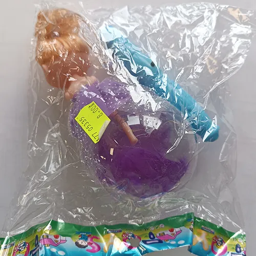 Poupée en plastique dans une robe textile violette. Les pattes de la poupée sont placées dans une boule en plastique transparent avec des plumes. Un contrôleur, y compris une batterie, est fixé à la tête de la poupée.